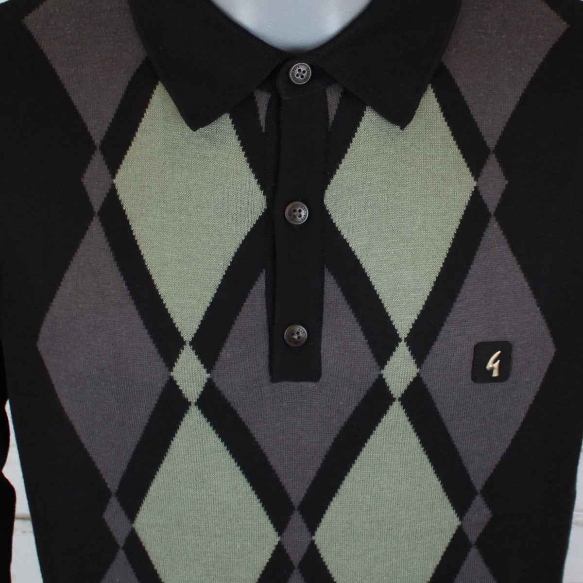 A close-up of a knit polo sweater with an argyle pattern in black, grey, and sage green, featuring a black collar and three-button placket.