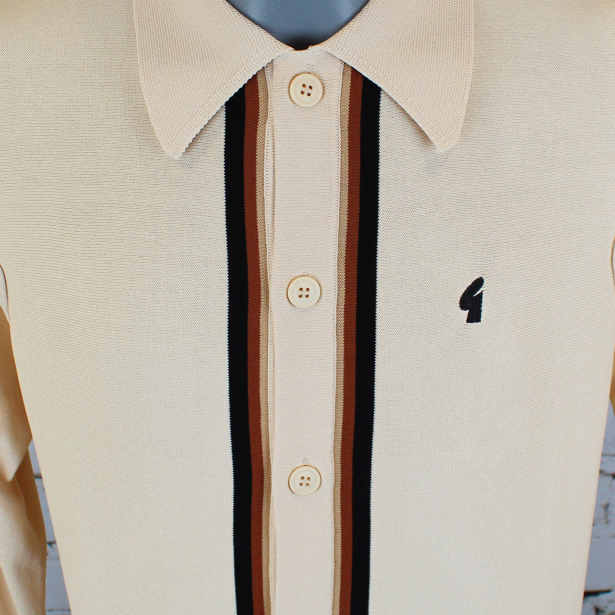 Close-up of a cream-colored knit polo shirt on a mannequin, featuring vertical stripes down the center in black, brown, and rust, with a small black embroidered logo on the chest.