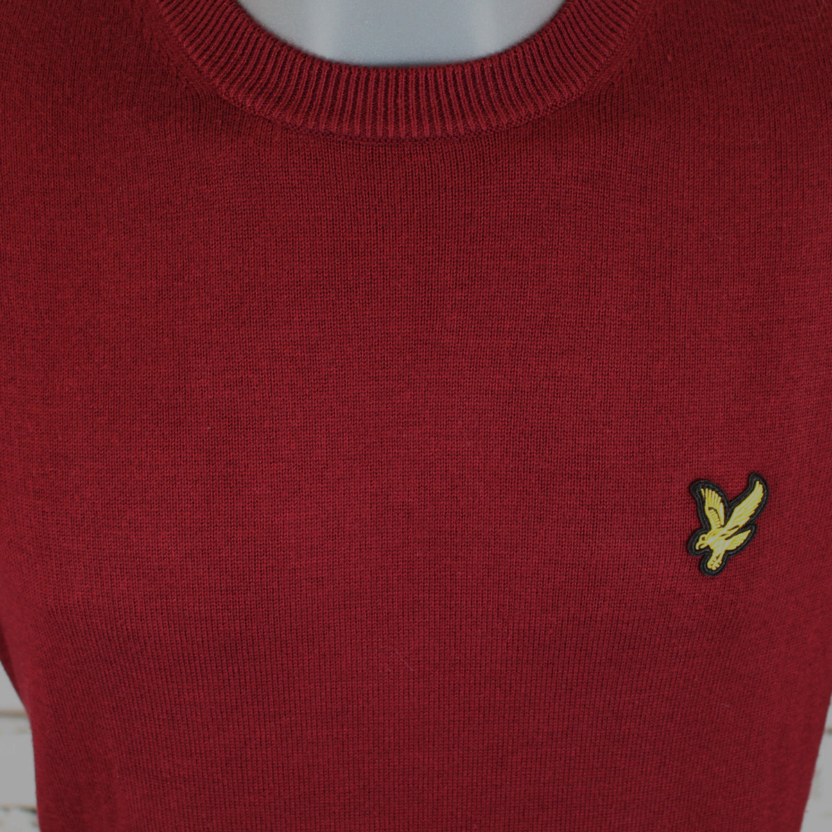 A close-up of a dark red ribbed knit sweater on a mannequin, featuring the embroidered yellow Lyle's bird logo on the left chest.