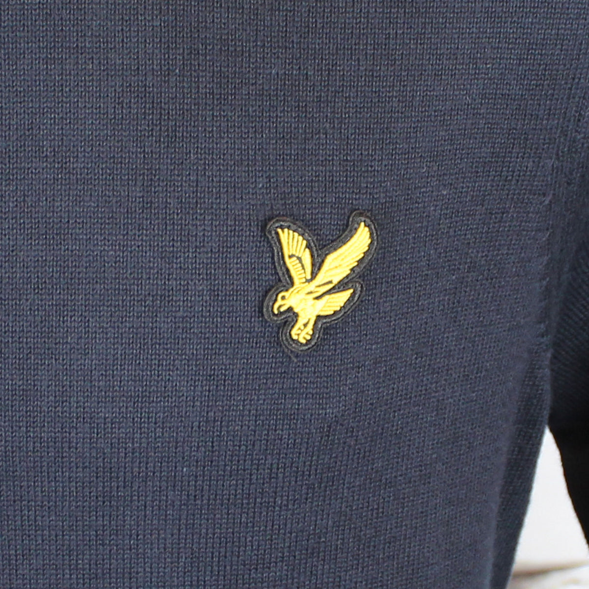 Close-up of a navy blue ribbed knit garment featuring a yellow embroidered Lyle's eagle logo patch on the chest.