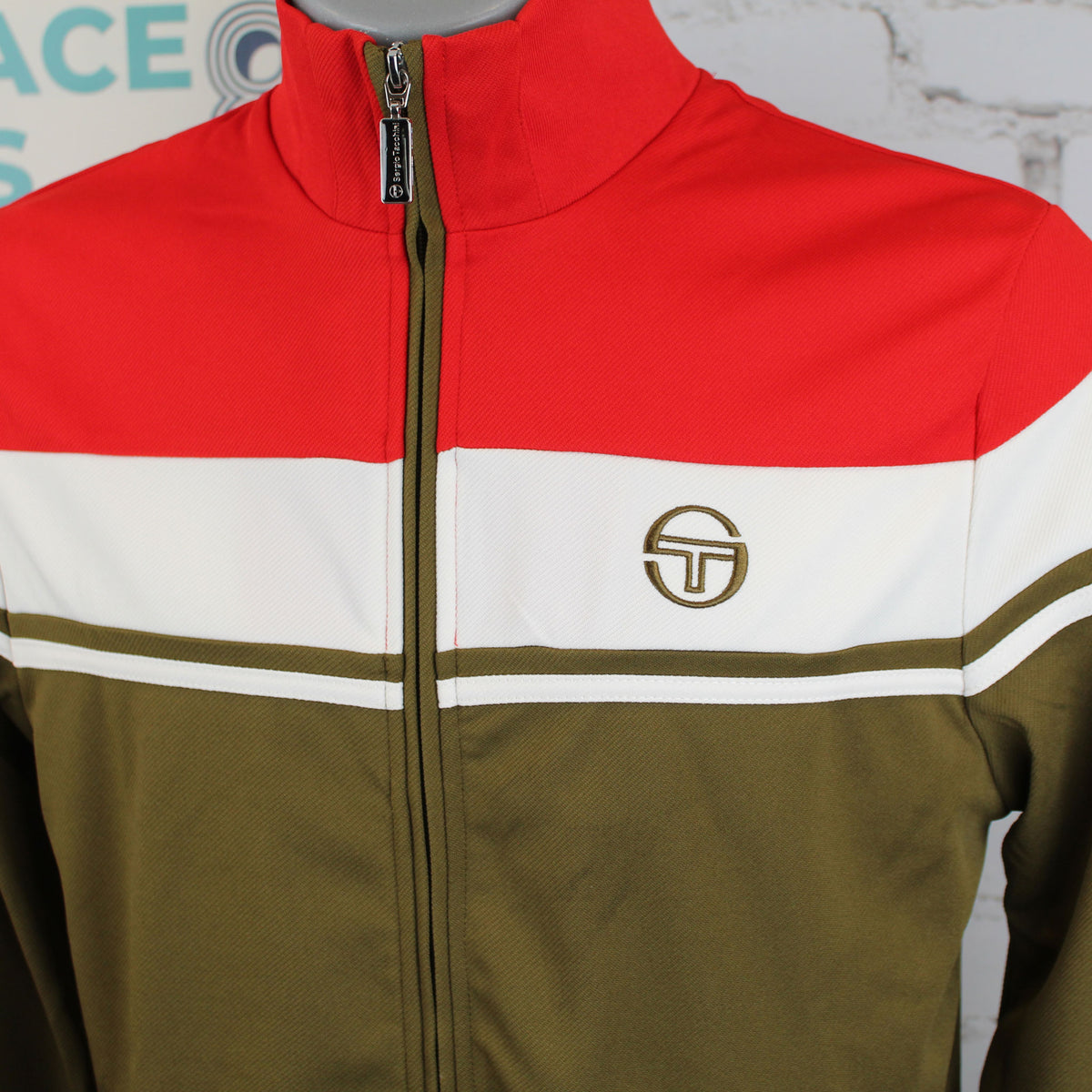 A close-up of a color-blocked zip-up jacket on a mannequin torso. The jacket features a bright red upper chest and shoulders, a wide white horizontal stripe across the chest, and an olive green lower body and zipper trim.