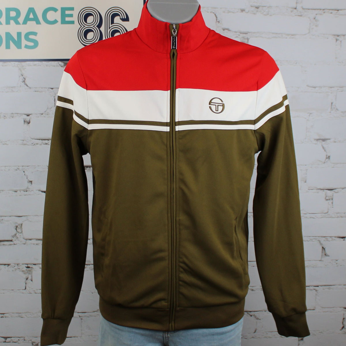 A vintage-style zip-up track jacket displayed on a mannequin torso against a white brick wall. The jacket features a red yoke, a white stripe with olive green double lines across the chest, and an olive green body and sleeves.