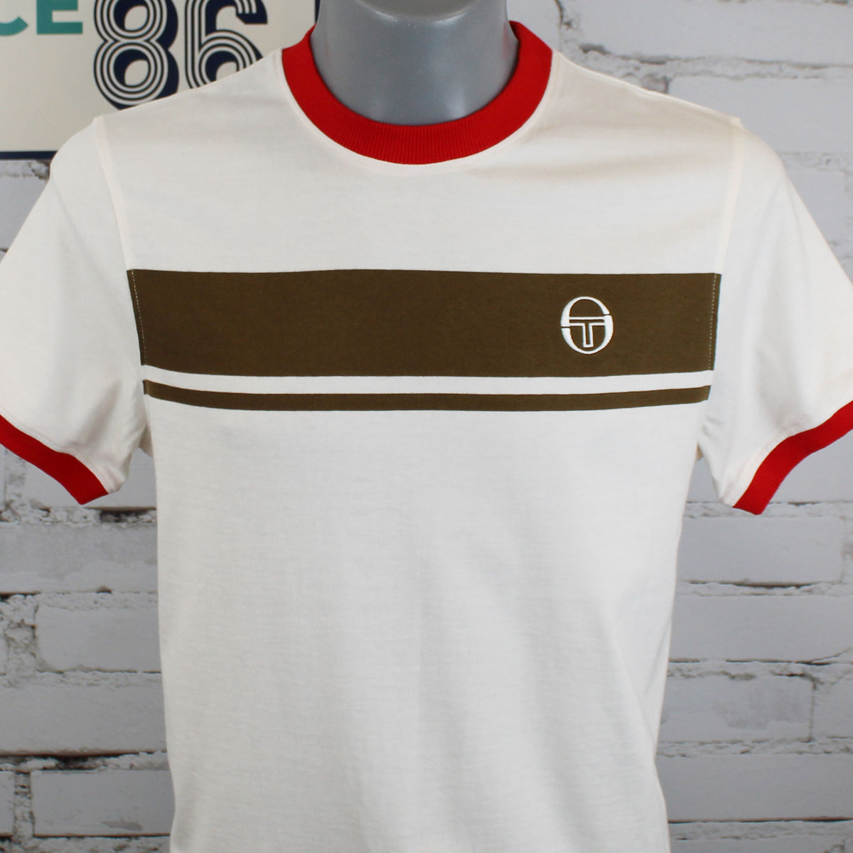 A vintage-style short-sleeve t-shirt displayed on a gray mannequin torso against a white brick wall. The shirt is off-white with a bold brown horizontal chest stripe, a thin white line through the middle, red collar and sleeve trim, and a small circular monogram logo on the left chest.