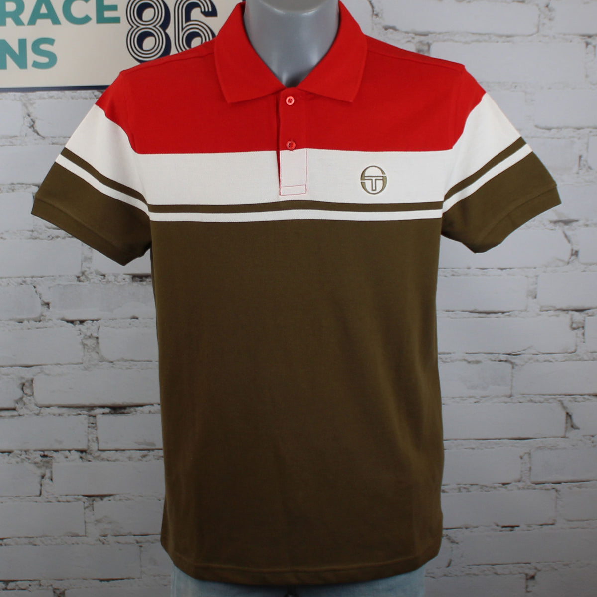 A short-sleeve polo shirt displayed on a mannequin torso against a white brick wall. The shirt has a color-blocked design with a red yoke, a wide white stripe, and a dark brown lower body and sleeves.